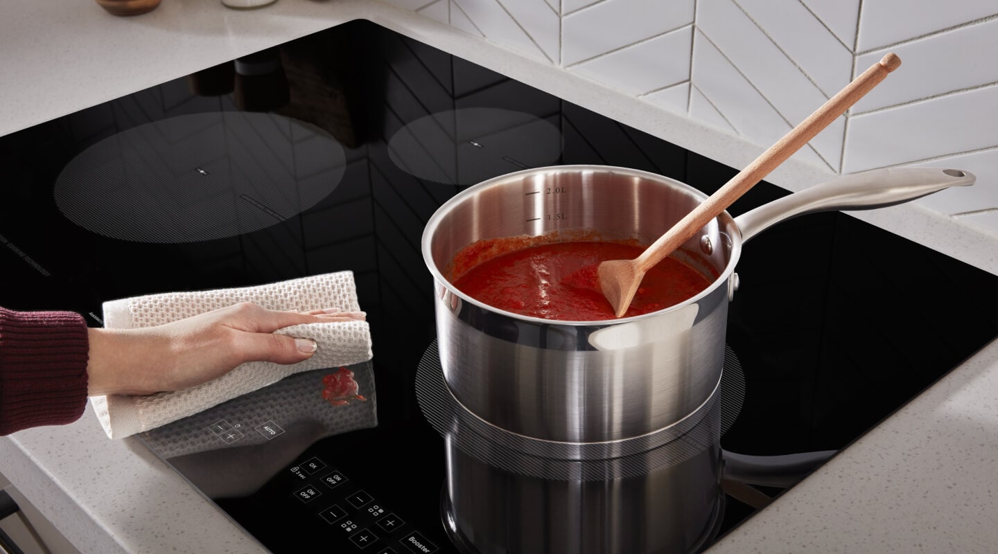 Person wiping cooktop next to burner with saucepan Person wiping cooktop next to burner with saucepan