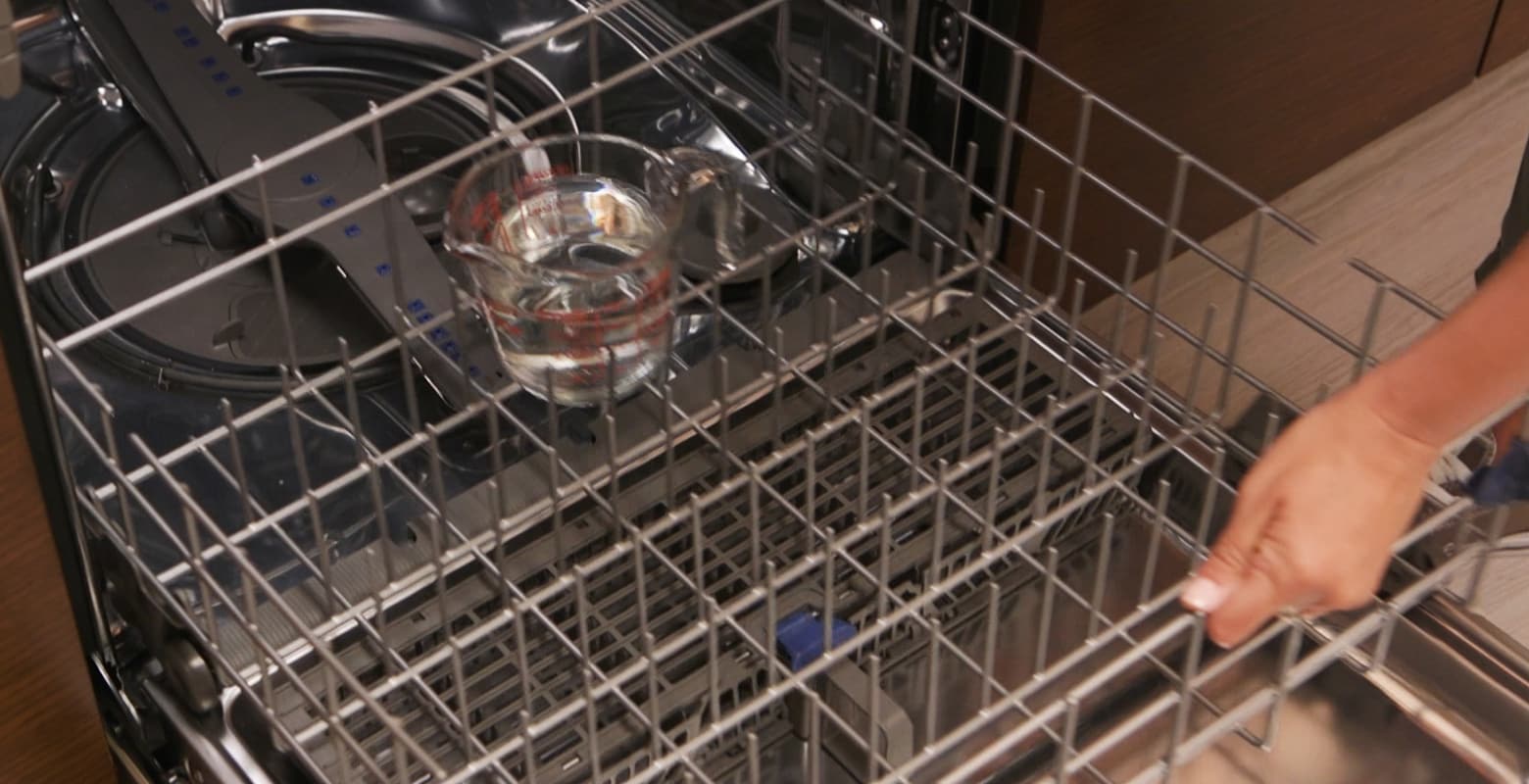 A person adding a measuring cup of vinegar to a dishwasher rack