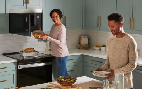 A couple making dinner in a sea green kitchen with one of them pulling a casserole dish out of a microwave