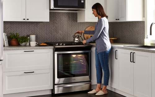 Person standing over a gas range with a hand on a tea kettle that’s resting on a stovetop in a kitchen with white cabinetry