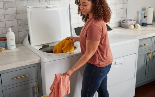 A person loading orange and pink linens into a white top load washer next to a white front load dryer