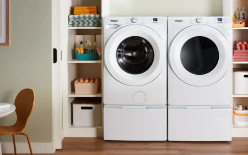 A white Whirlpool Front Load Washer & Dryer pair in a laundry closet with shelves of laundry supplies