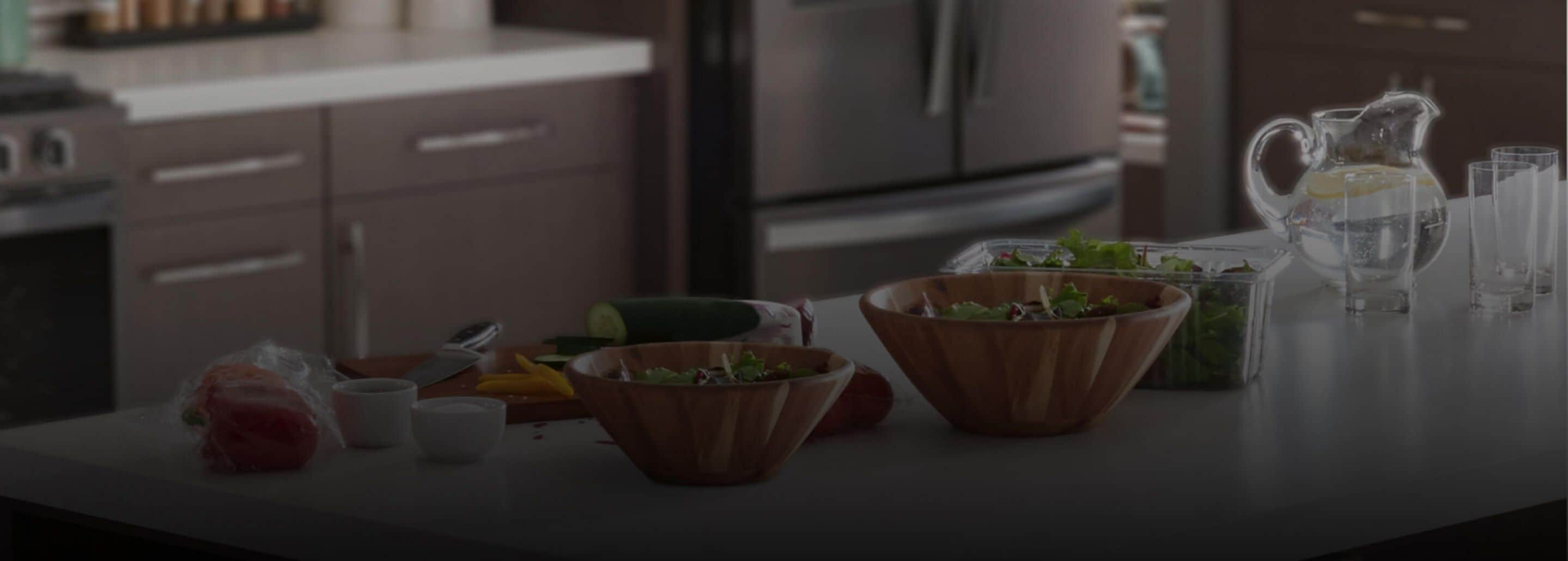 A contemporary kitchen island featuring two wooden salad bowls surrounded by various ingredients, plus a pitcher of clean, clear water.